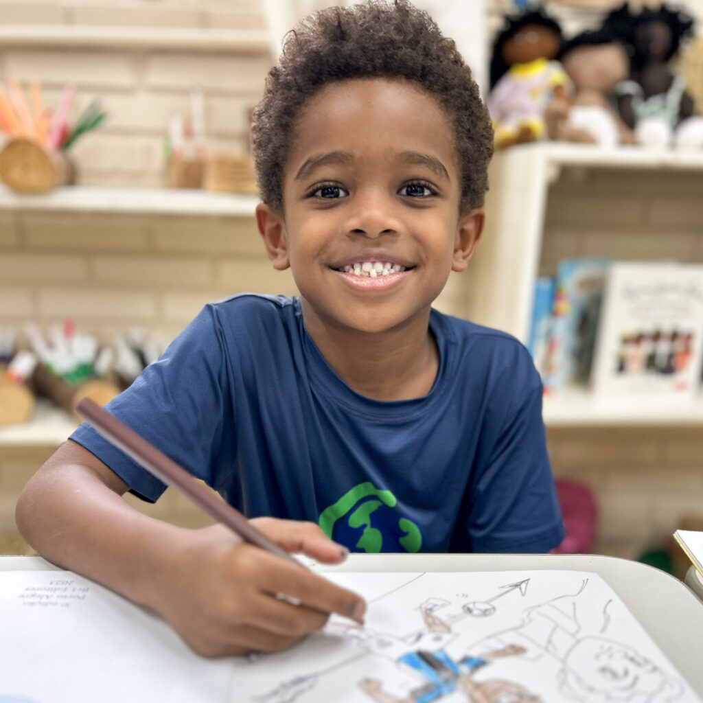 Menino negro sorrindo para a câmera. Em sua mão está um lápis de cor marrom. Ele está colorindo um desenho.