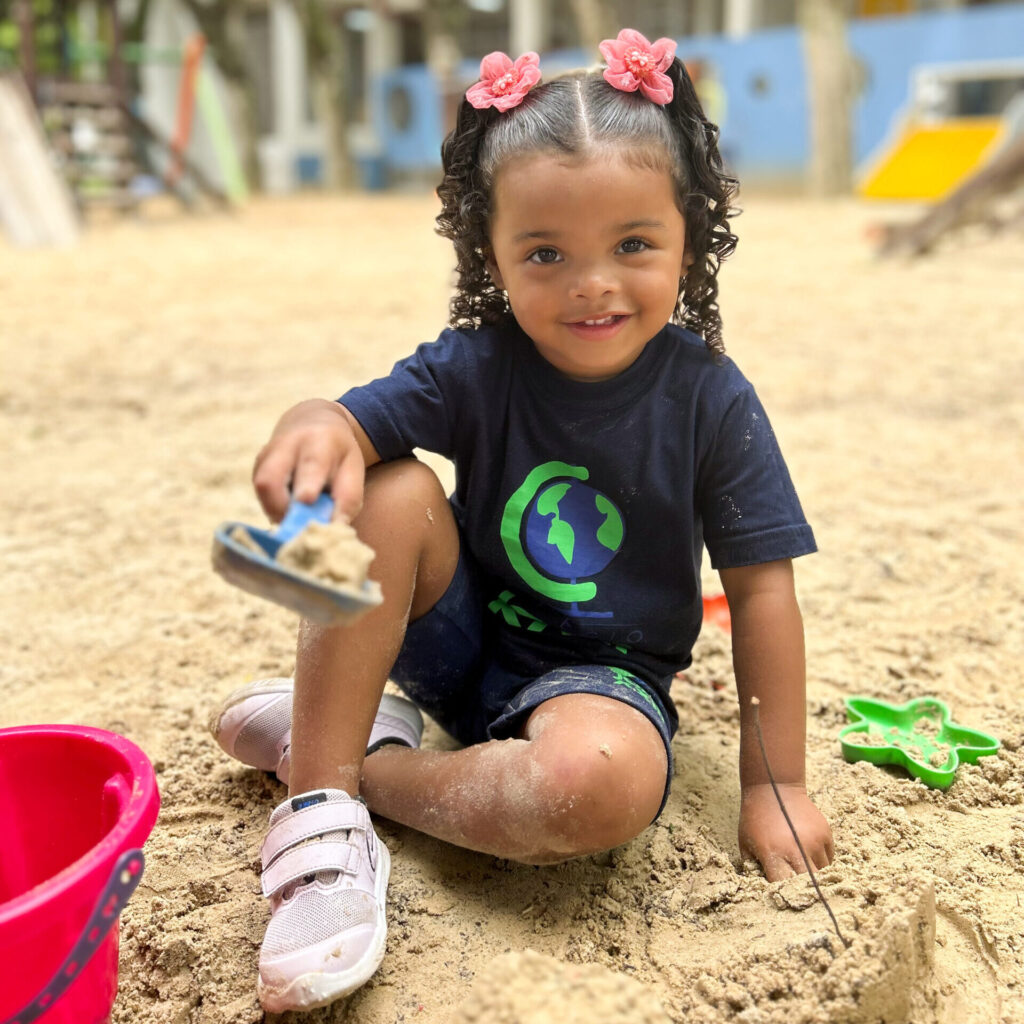 Menina sorrindo para a câmera. Ela está sentada no chão de areia, envolta de brinquedos. Em sua mão ela segura pazinha com areia.